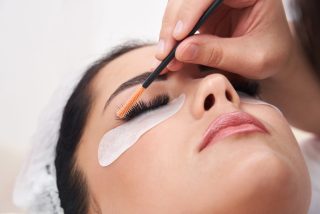 Cosmetologist making eyelash extention and correction using brush in salon